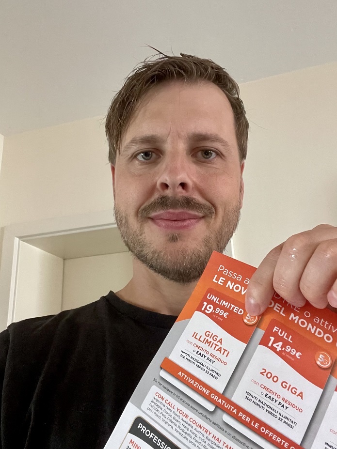 Tobi mit dem Papierflyer von WindTRE. Er zeigt auf das genutzte Prepaid Angebot für 19,99 € pro Monat mit unbegrenztem Datenvolumen in bis zu 5G.