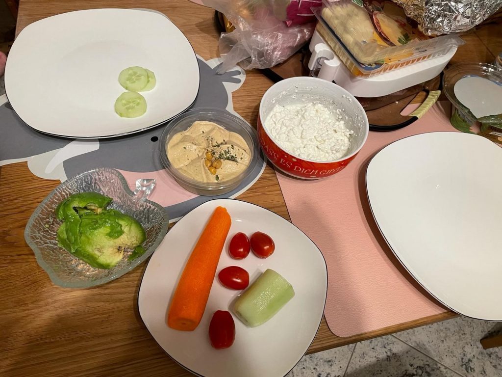Das Bild zeigt im Kontext Essen und Ernährung - Das Abendessen später im Text weiter beschriebenen Nahrungsmittel, im Vordergrund ein Stück geschälte Salatgurke, eine geschälte Karotte, 4 kleine Tomaten, Hummus, eine geschälte Avocado und körnigen Frischkäse in einer Schüssel.